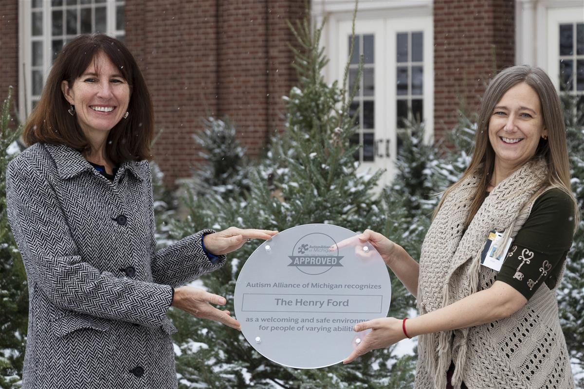 Two women hold a frosted plastic plaque with text in front of snow-covered evergreens and a brick building