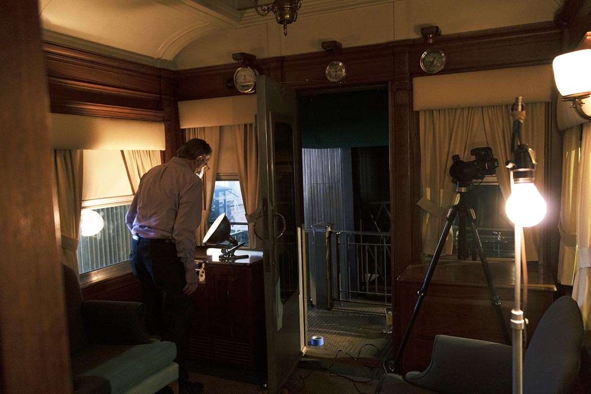 Photographer Rudy Ruzicska, setting up lights for our first interior shot of the Fair Lane railroad car lounge Interior space with coved ceiling, containing a man bending over a light, with other lights and a camera on a tripod in the space