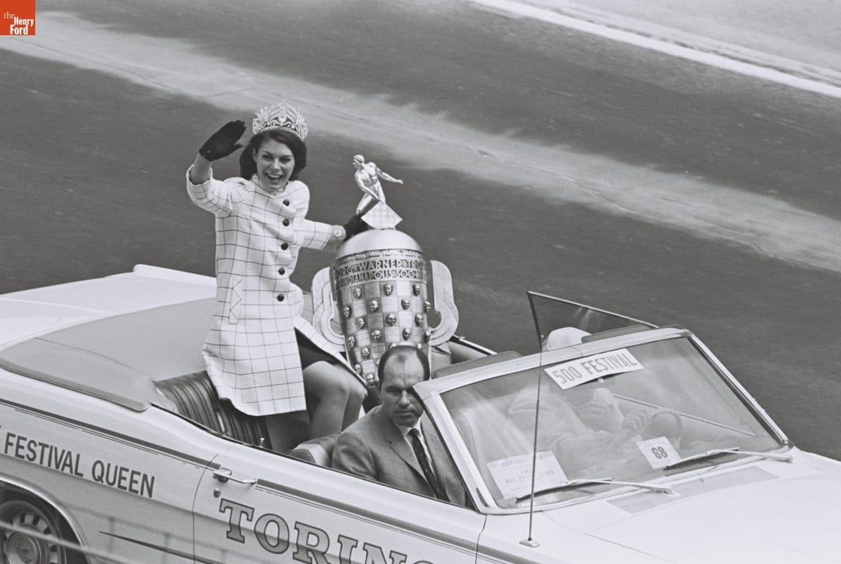 Woman wearing checked coat, tiara, and gloves and holding a very large trophy sits on the back of an open convertible and waves, with two men in the front seats