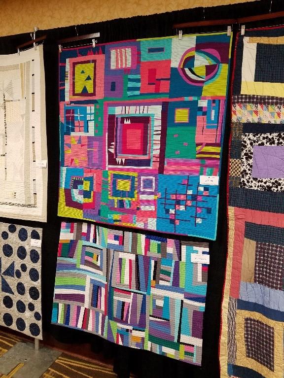Quilt display at Lincoln, Nebraska Modern Quilt Guild at American Quilt Study Group’s October 2019 Seminar Brightly colored quilts hanging on a black backboard