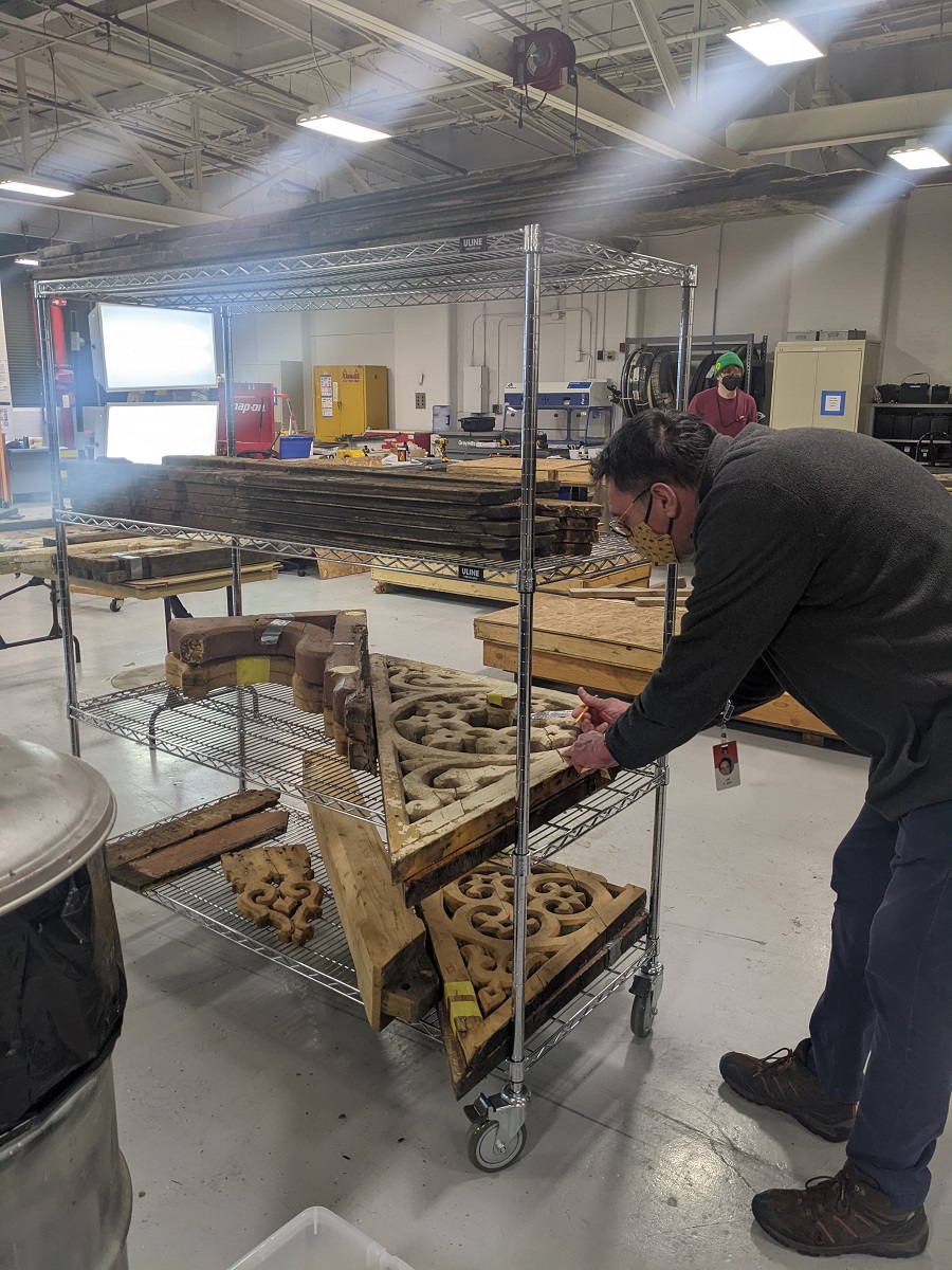Man leans over wooden architectural elements on metal shelving in a large, sunny warehouse space