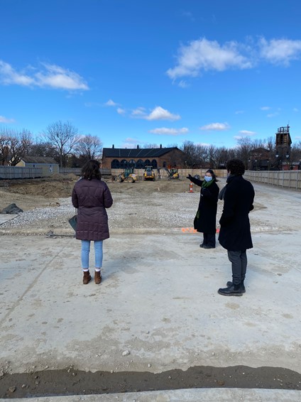 Two people look at a construction site, while a third in a mask looks at the first two and gestures toward construction site