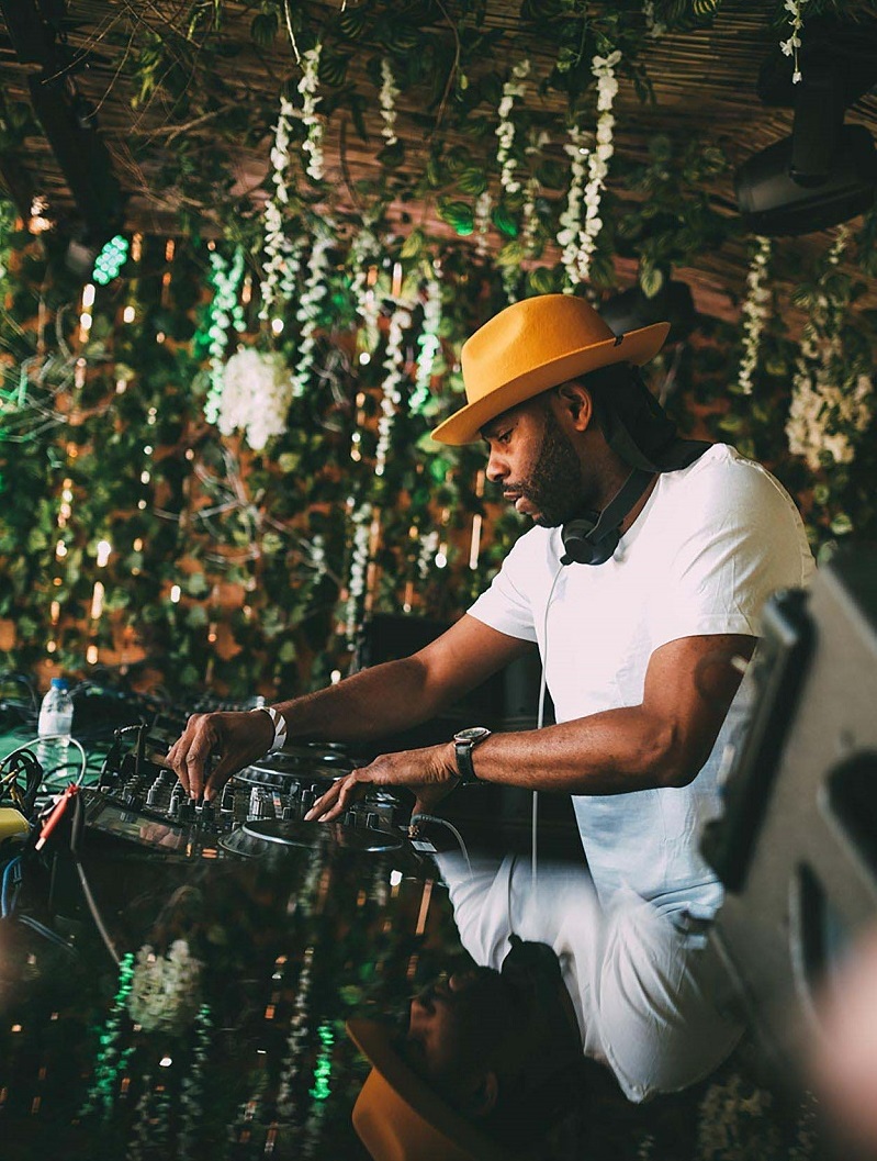 Waajeed performing at Brunch Electronik Lisboa in Portugal African American man in white t-shirt and yellow hat works at a DJ mixing station with foliage descending from wooden walls and ceiling in the background