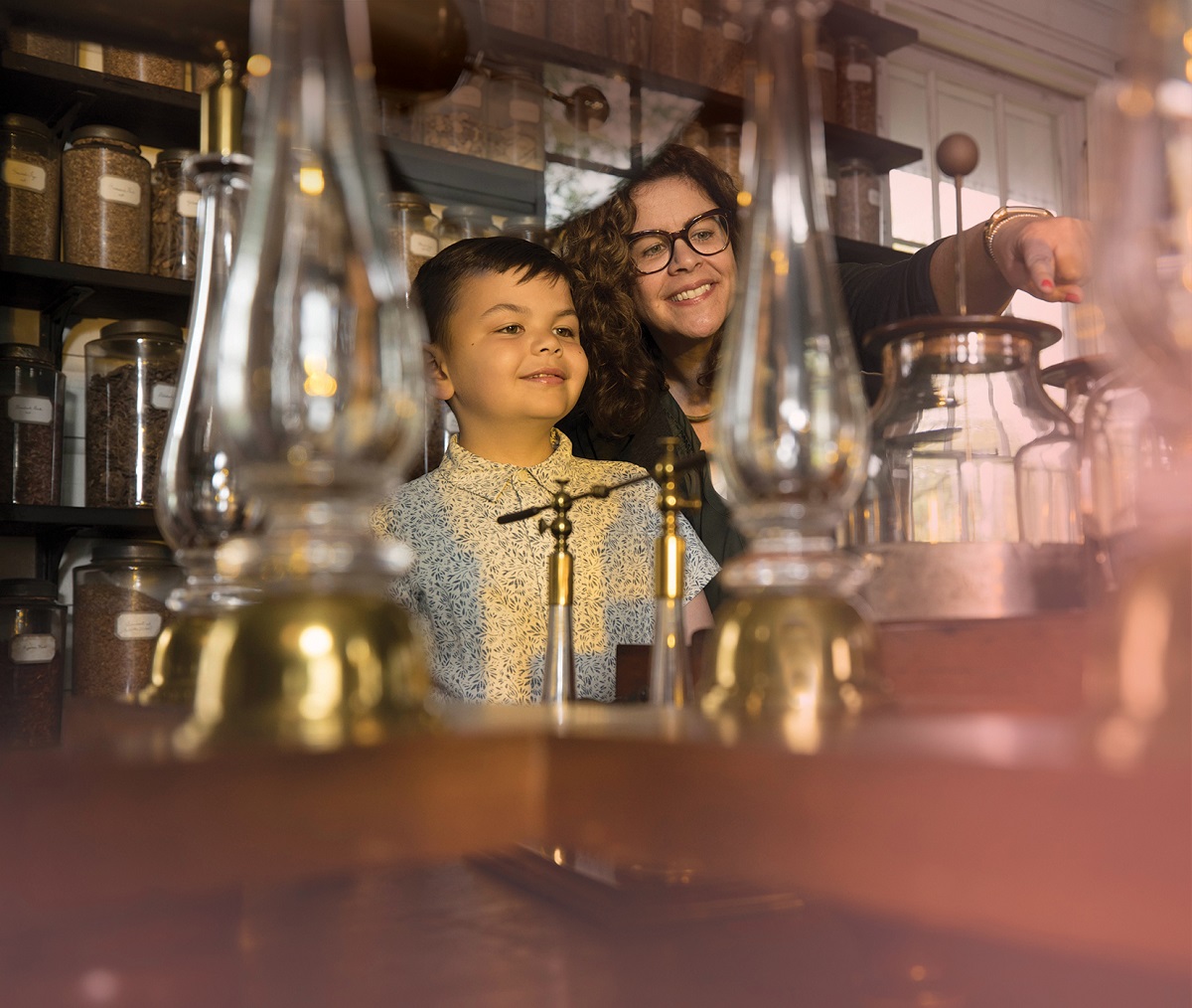 A woman and young boy smile, point, and look at a table filled with glass jars and lamps, with shelves of more glass jars behind them