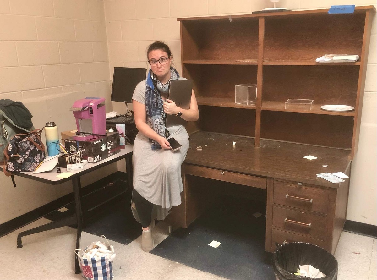 Frowning woman sits on edge of desk with hutch next to table filled with bags and boxes