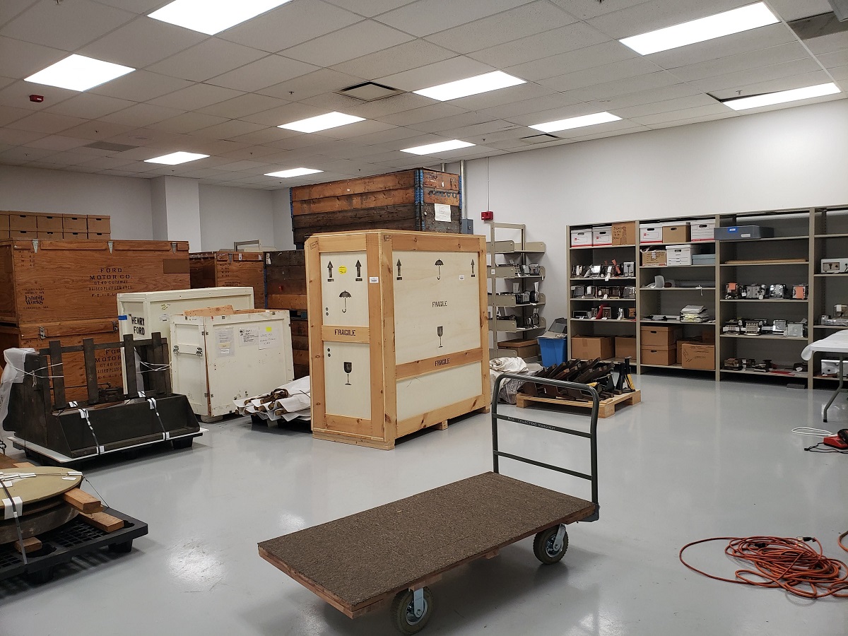 Room containing crates, shelving full of items, and large items on pallets