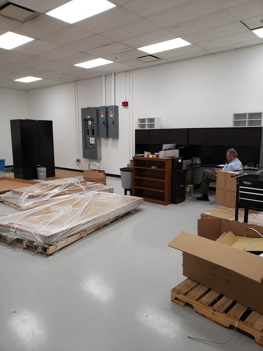 Man sits at empty desk in room containing a few shrinkwrapped piles of wood, pallets, and empty furniture