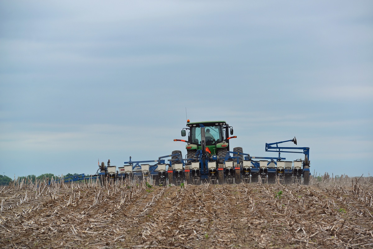 A green tractor trailing a long blue machine hitched behind it travels through a field of dirt and stubble