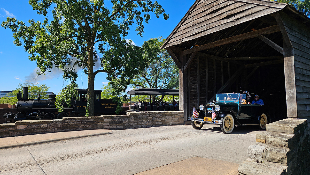 A Model A car driving through a covered bridge with a black train in the background