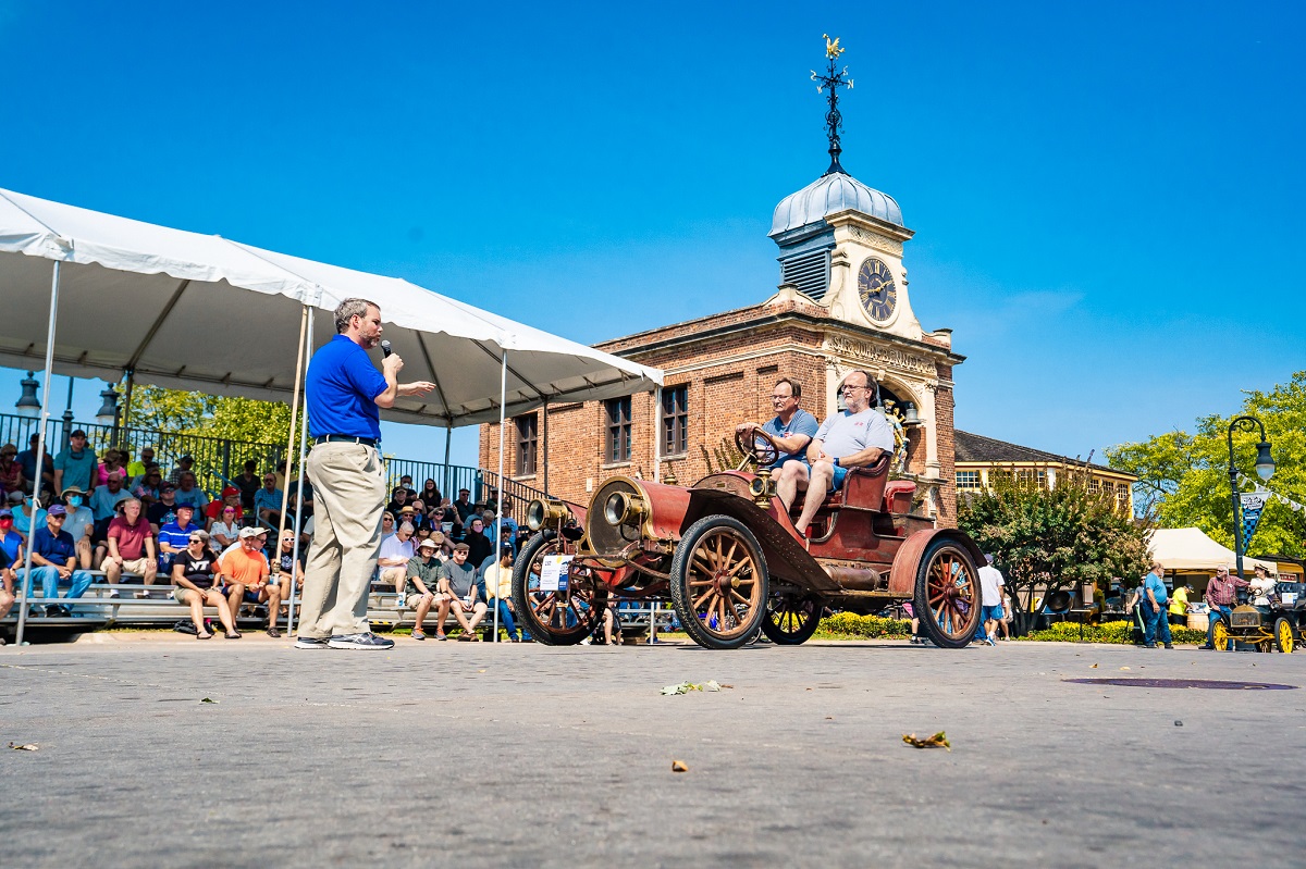 Pass-in-Review at Old Car Festival 2021 Man in a blue shirt and khakis holding a microphone gestures to an old-fashioned open car with two people in it, as spectators look on from the side