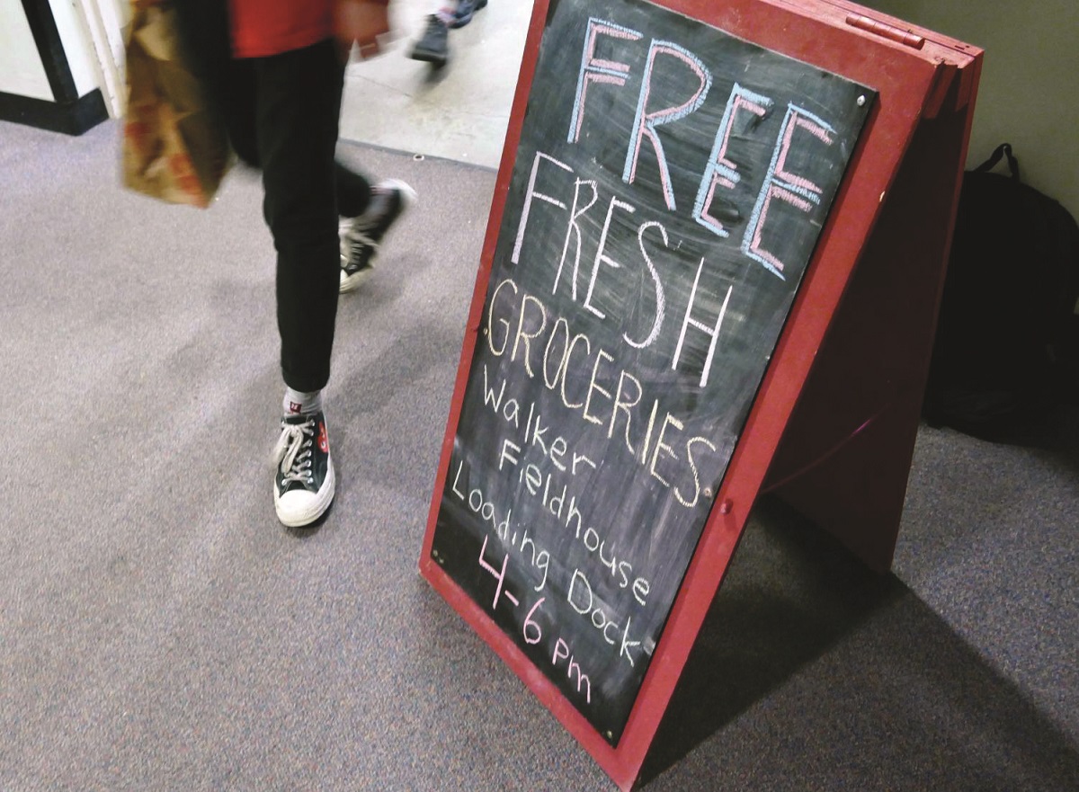 "Free Fresh Groceries" sign Standee-type blackboard sign reading "Free fresh groceries: Walker Fieldhouse Loading Dock 4-6 PM" with person walking by it holding a paper grocery bag