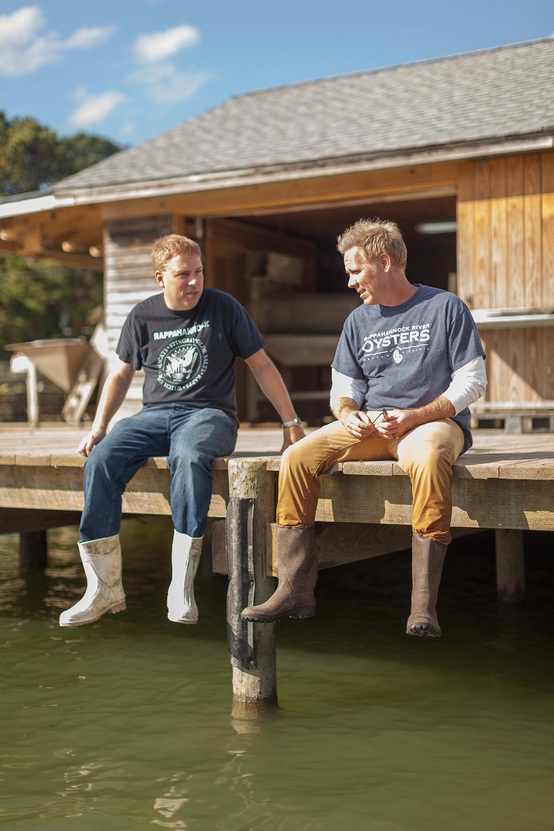 Two men wearing boots, khakis/jeans, and t-shirts sit with their legs dangling off a dock with a wooden building in the background 