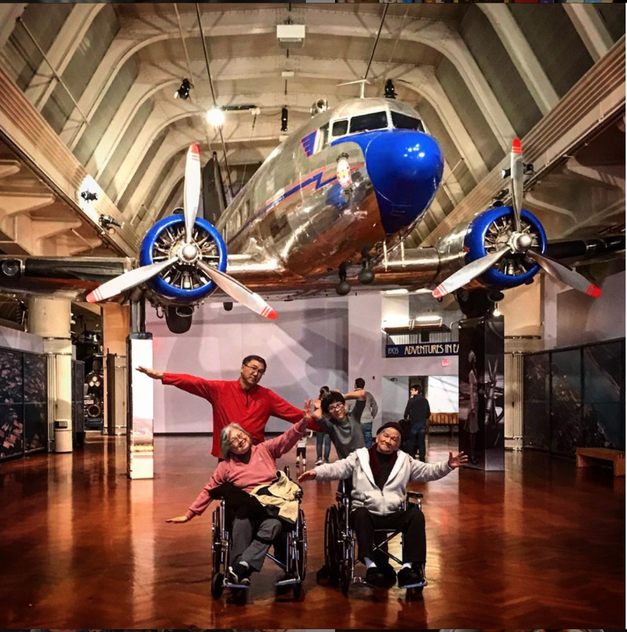 Four people (two in wheelchairs) pose with arms outstretched underneath a silver-and-blue airplane