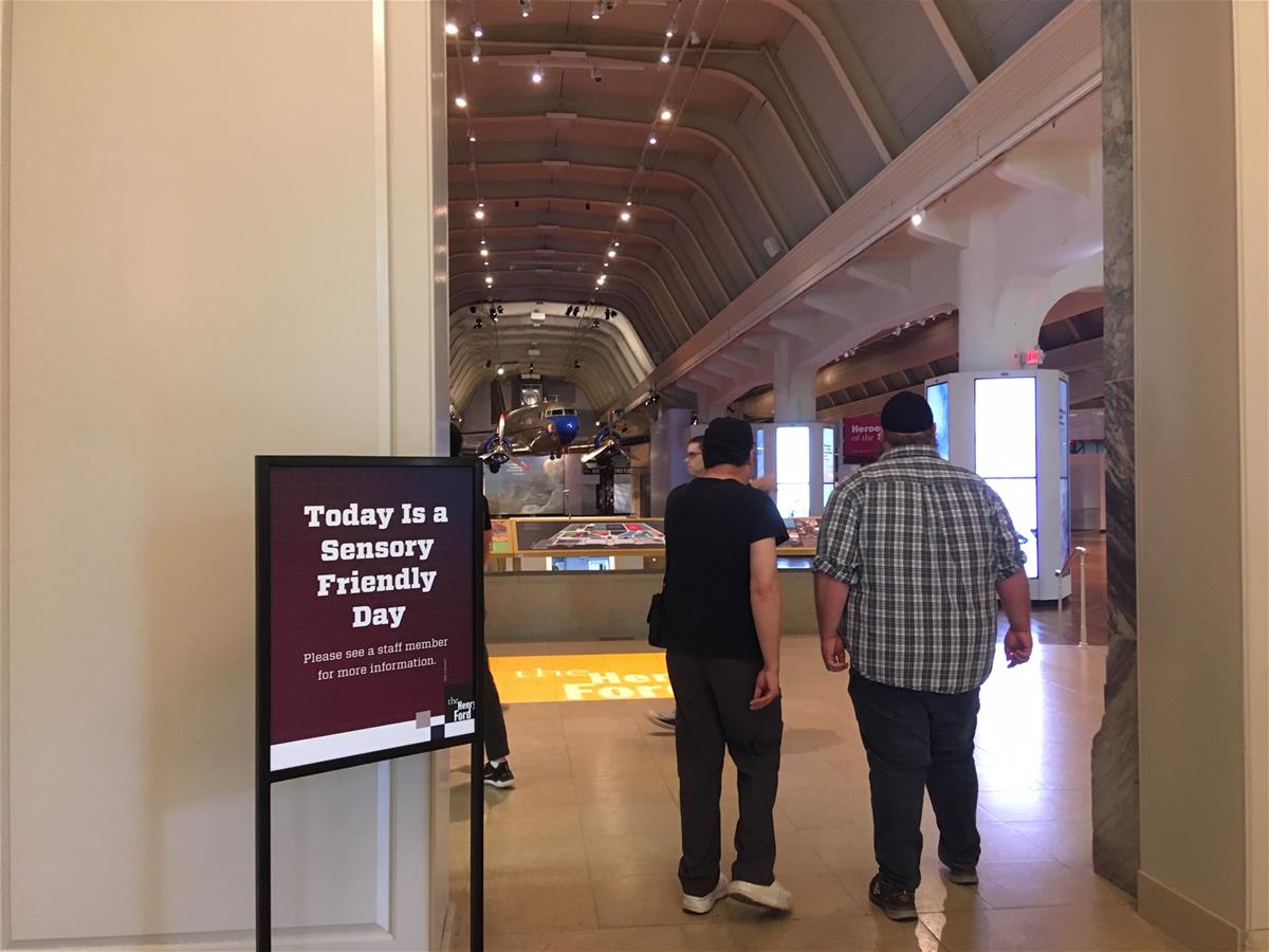 Two people walk past a sign with text into a large high room with an airplane hanging from the ceiling 