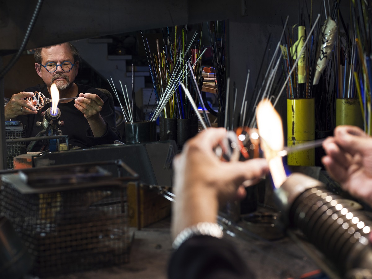 Man in workshop holds glass in a flame, seen in a reflection in a mirror on the wall