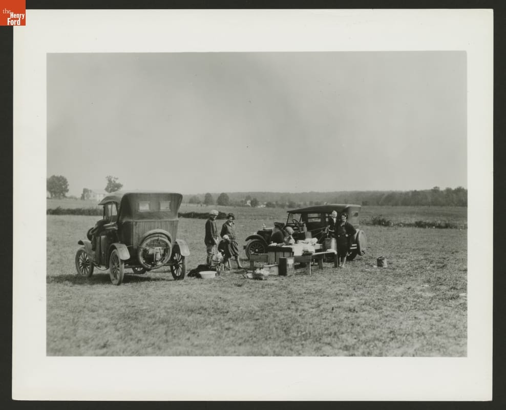 Small group of people in a field with two cars and a table covered in gear