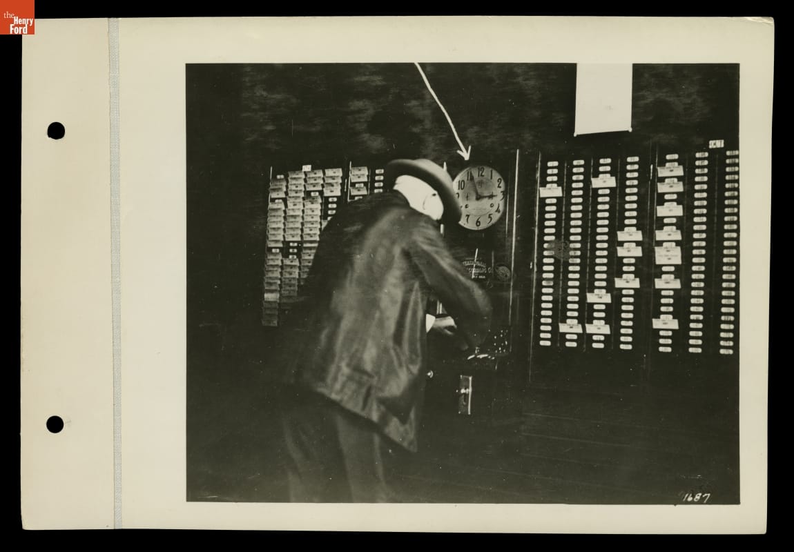 Thomas Edison Punching the Time Clock at His West Orange, New Jersey Laboratory, August 27, 1912 Man in suit and hat slightly leaning over towards a clock and wall with many slots for cards