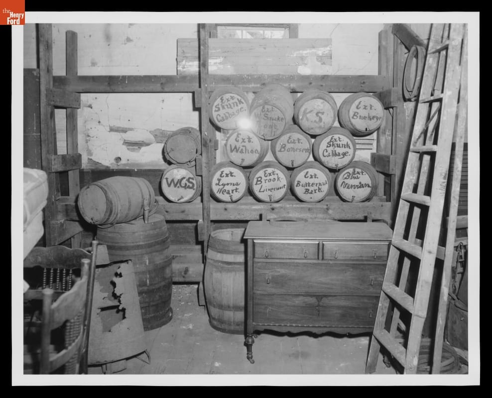 Black-and-white photo of storage room containing casks on racks, barrels, and furniture