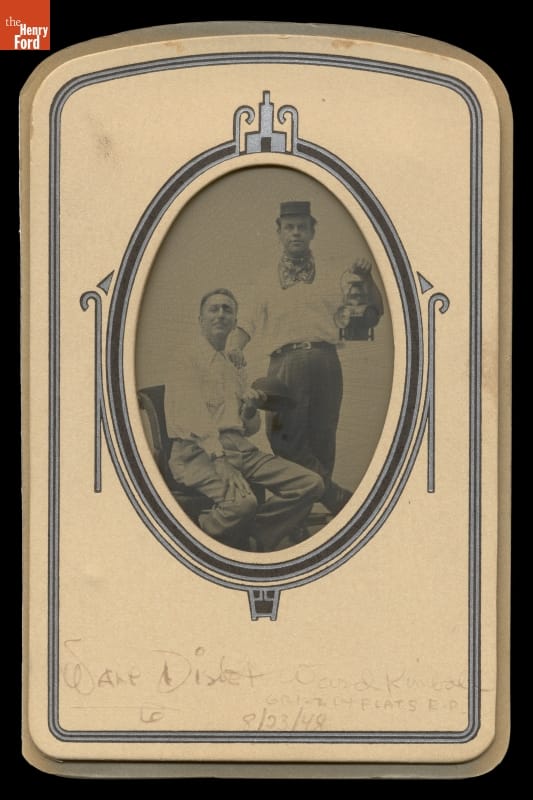 Two men, standing one holding a lantern and seated one holding a helmet, pose for a black-and-white photo