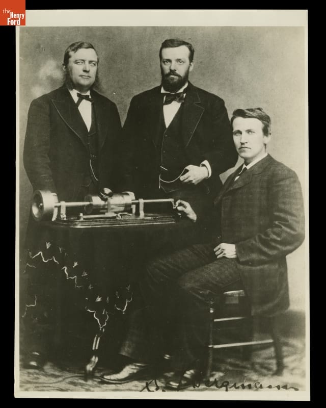 Three men in suits, two standing and one seated, around a piece of equipment