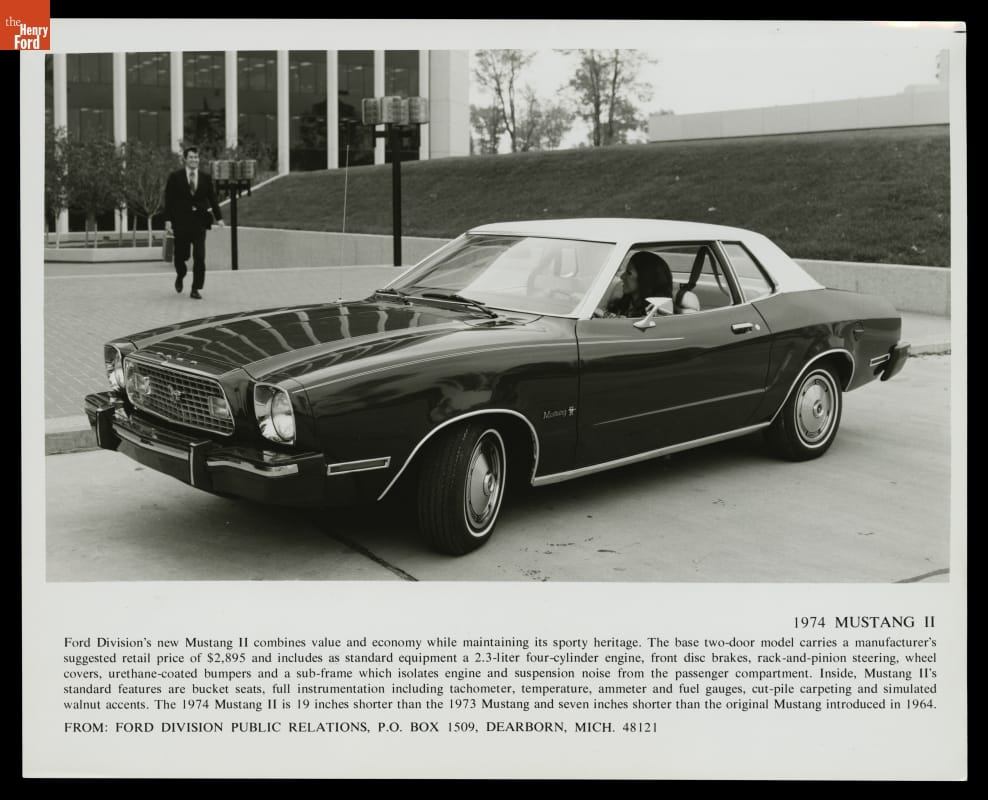 Black-and-white photo of car in front of building with concrete plaza and grass berm; one person is behind wheel and another in suit walks toward car