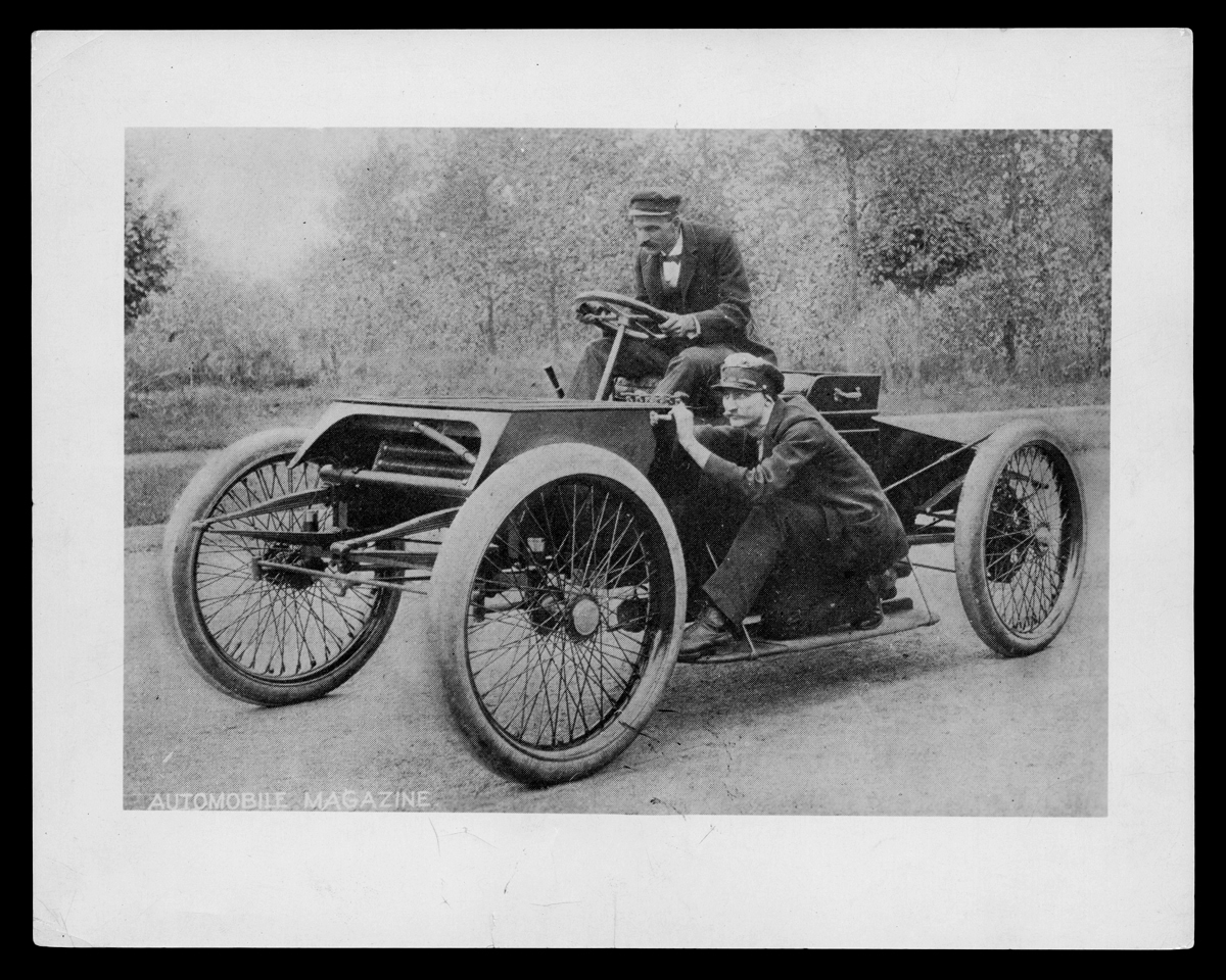 Man sits in early open race car, with another man crouching on running board