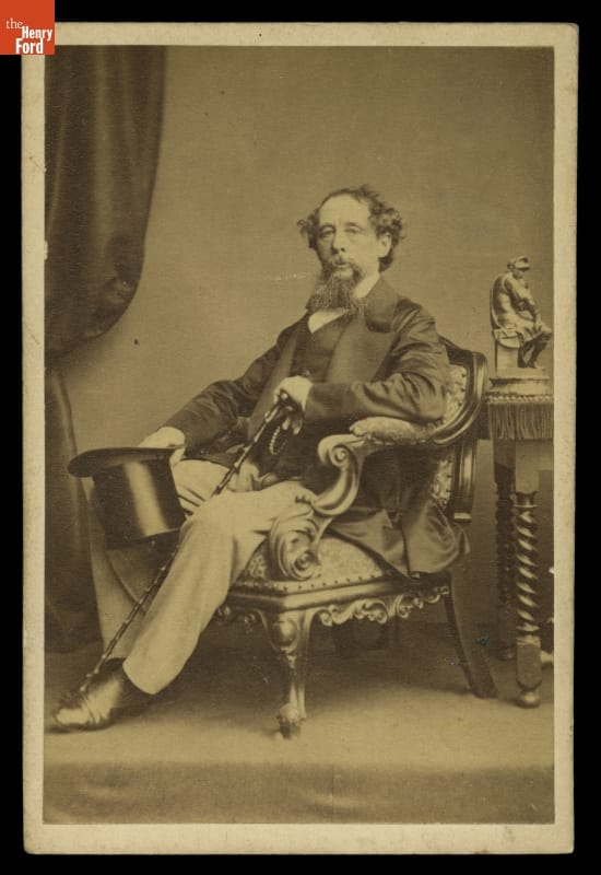 Portrait of Charles Dickens, 1865-1870 Man with beard and mustache seated in chair, holding cane and top hat, next to a table with a statue on it and drapery