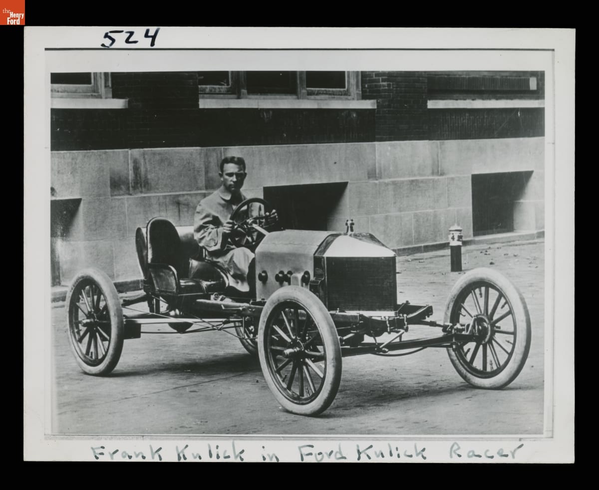 Black-and-white photo of man sitting in early open race car in front of building