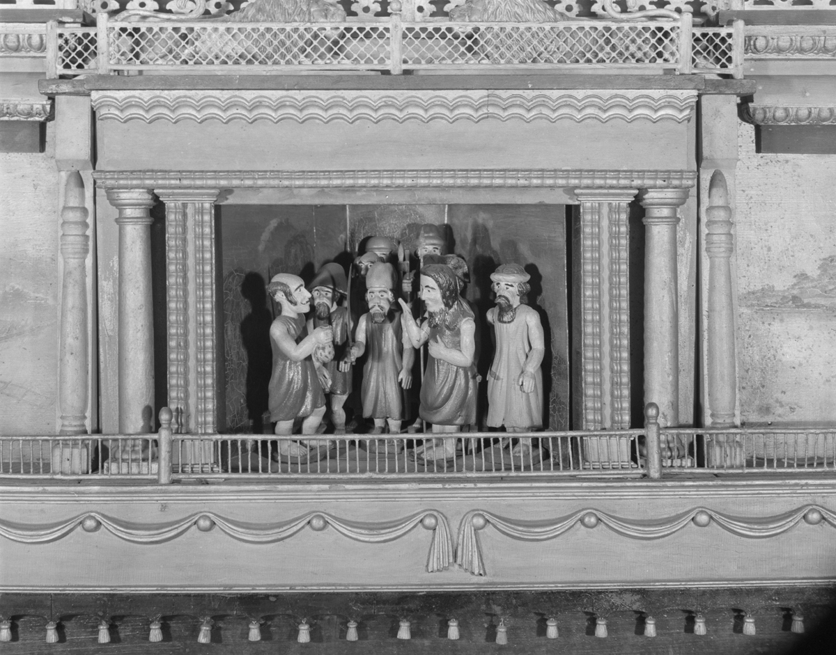 Black-and-white photo of scene with human figures wearing robes on a stage with carved details around them