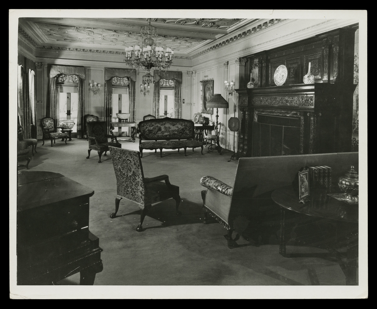 Black-and-white photograph of room with large, dark fireplace and ornate ceilings and furniture 