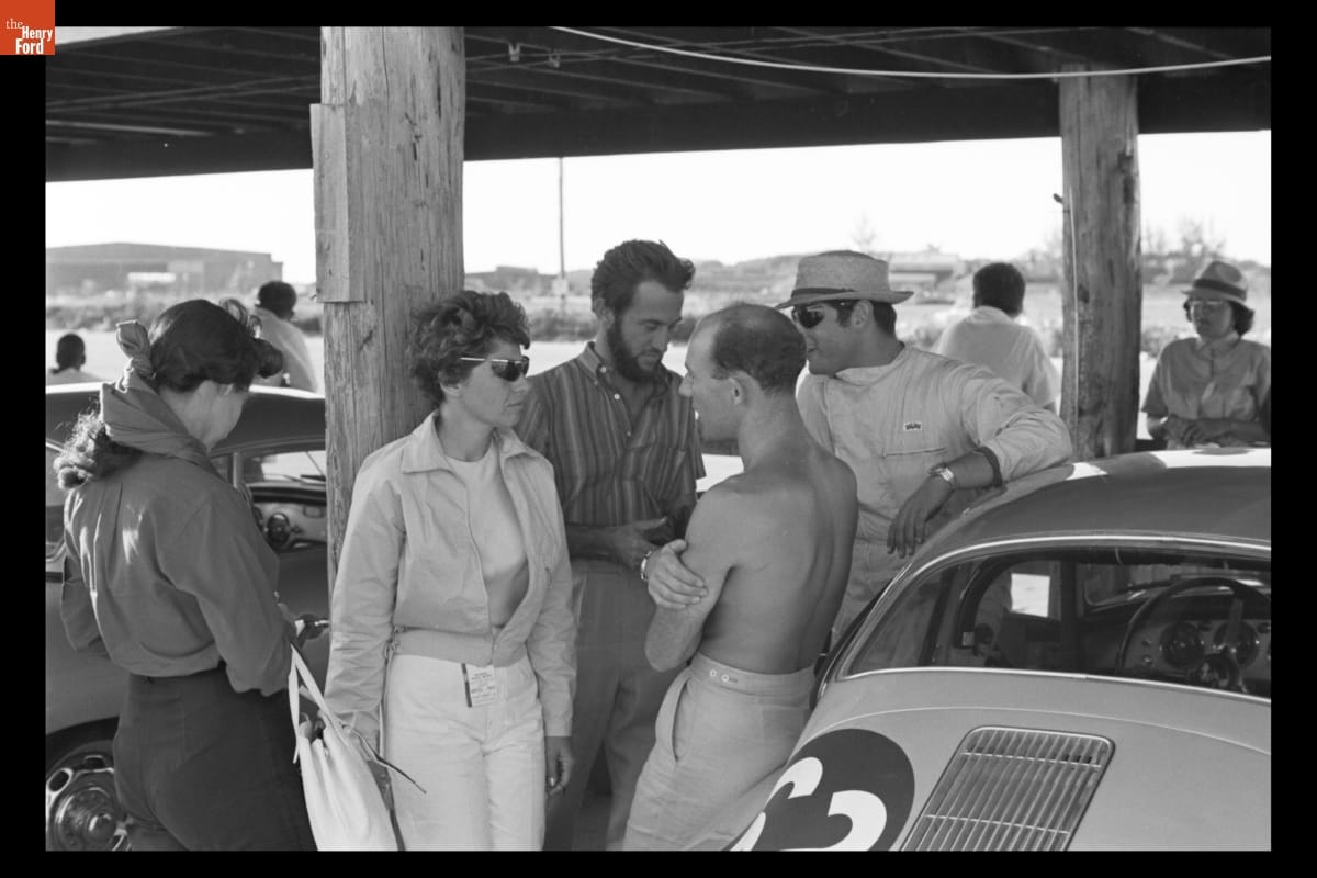 Woman leans against wooden post and talks to several other people, with additional people and cars nearby