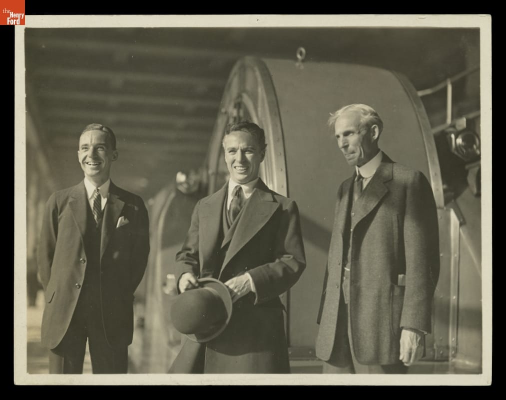 Three men in suits, one in middle holding hat, pose for a photo in front of large equipment or machinery