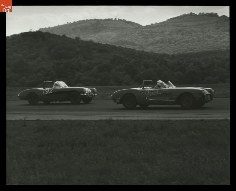 Two open-top race cars on a road or track with wooded hills in the background