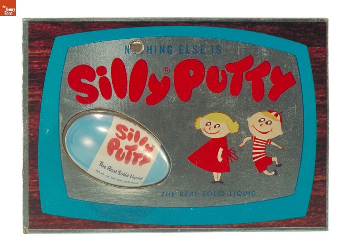 Plastic blister pack containing blue plastic egg with red and white label "Silly Putty" against blue, silver, and woodgrain background with text