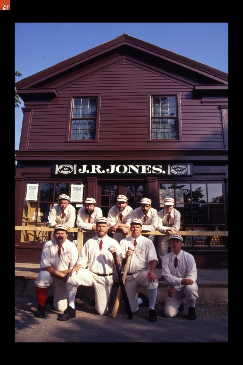 Nine people in old-fashioned baseball uniforms stand and kneel in front of a maroon wooden building with sign "J.R. Jones"