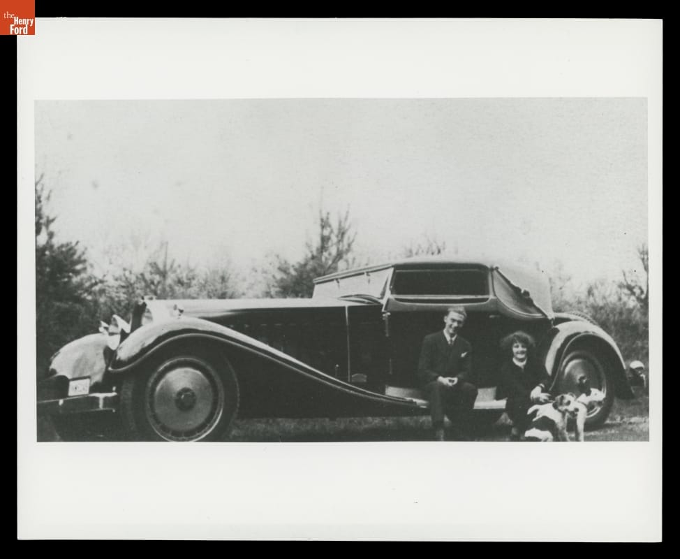 Black-and-white photo of a long car with swooping style lines; two people and two dogs sitting outside car