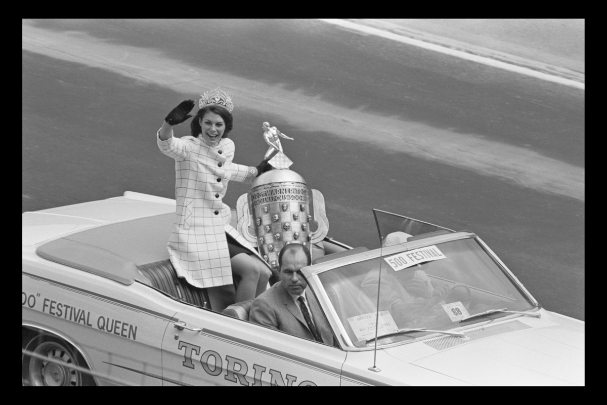 A woman waving in the backseat of a convertible with a large trophy
