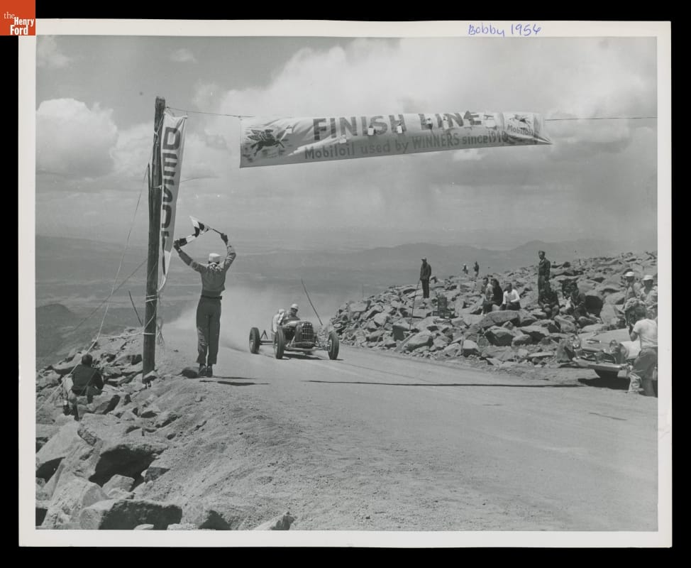 Car approaching banner marked "Finish Line" on dirt road with rocks on either side and steep dropoff on one side, mountains visible in background