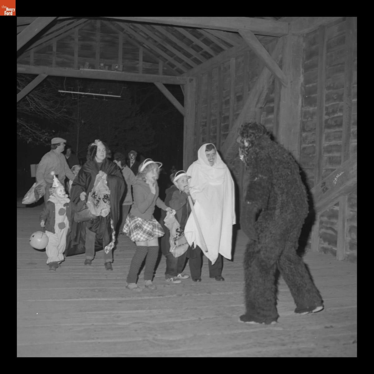 Person in a gorilla costume in front of adults and children, many in costume, in a wooden covered bridge