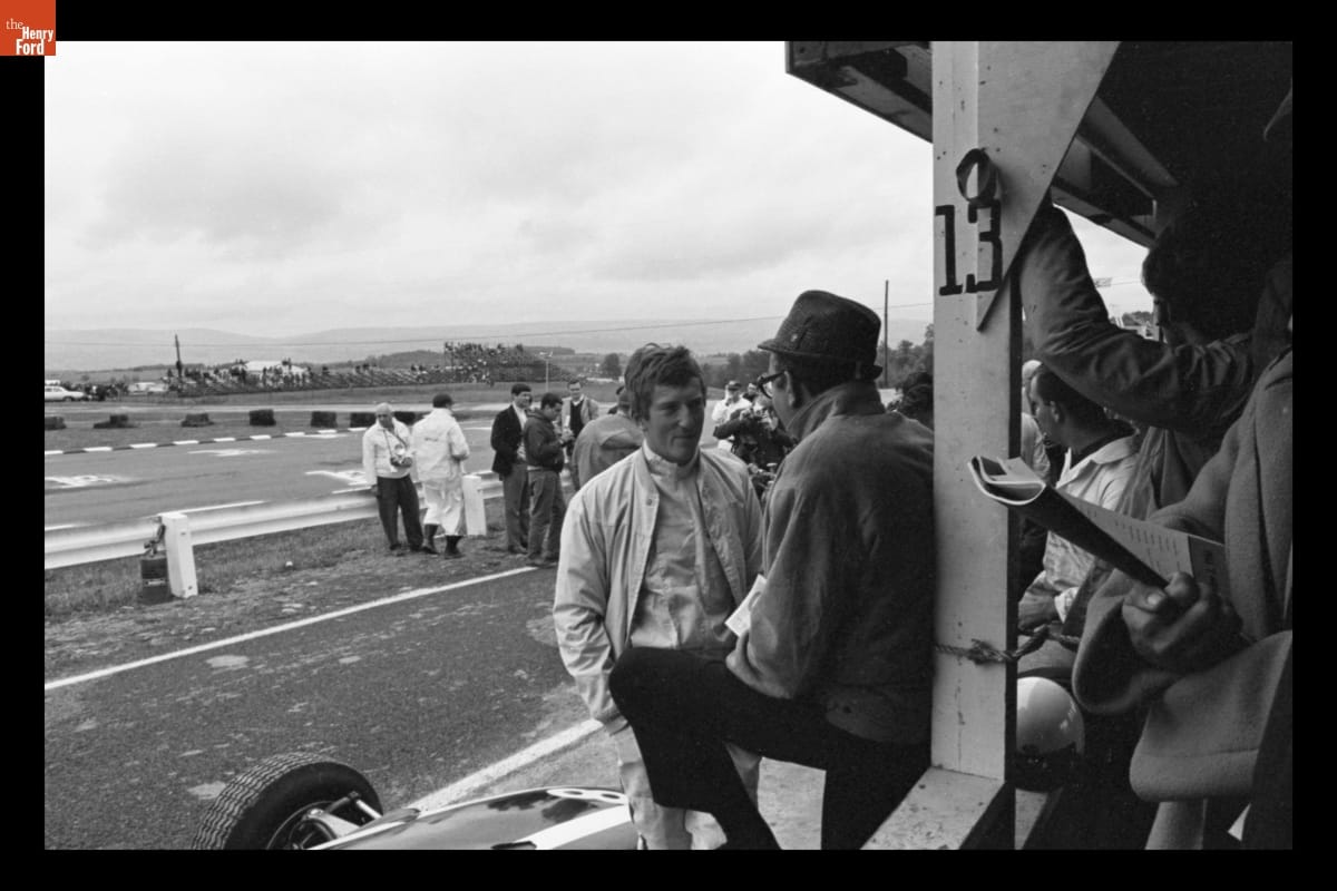 Jochen Rindt at the Grand Prix of the United States, Watkins Glen, October 1966 Two men talking; other people in foreground to right and in background are more people and a raceway