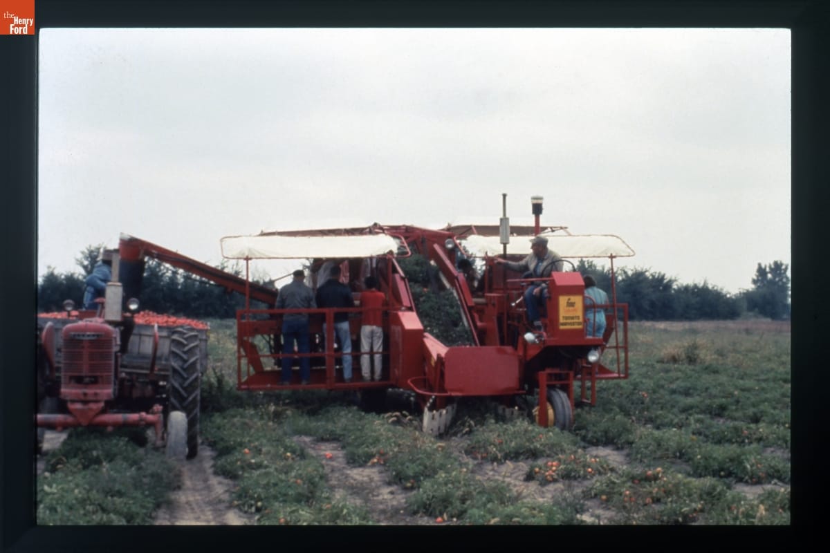 Large red wheeled piece of equipment in a field with a number of people riding on it
