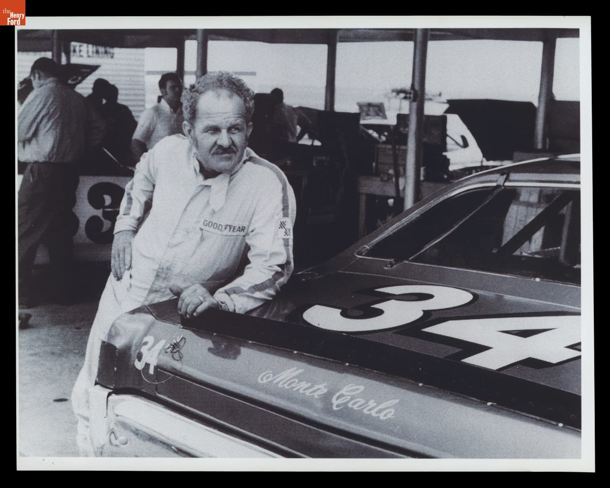 Wendell Scott with His 1971 Chevrolet Monte Carlo at the World 600, Charlotte Motor Speedway, May 24, 1972 Man in jumpsuit leans against trunk of racecar, with other people and cars in the background