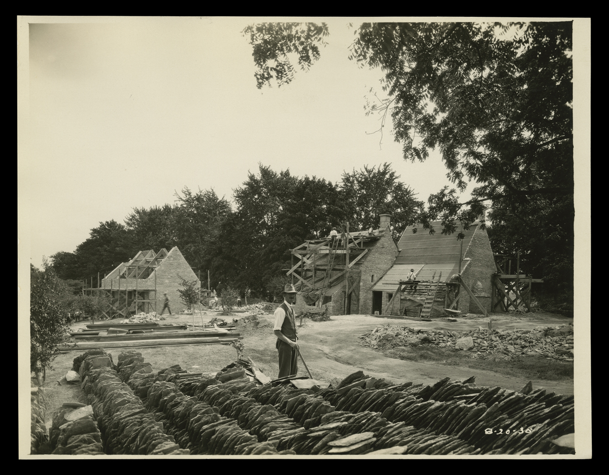 Rebuilding Cotswold Cottage in Greenfield Village, August 1930 Man stands next to rows of stacked shingles in foreground; two partially constructed buildings in background