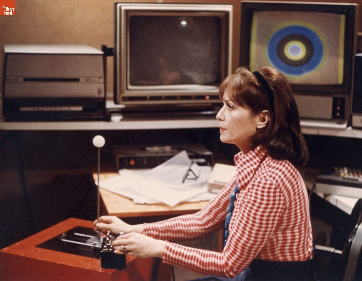 Woman in red-checked shirt surrounded by monitors and other computer peripherals manipulates a joystick device while looking straight ahead