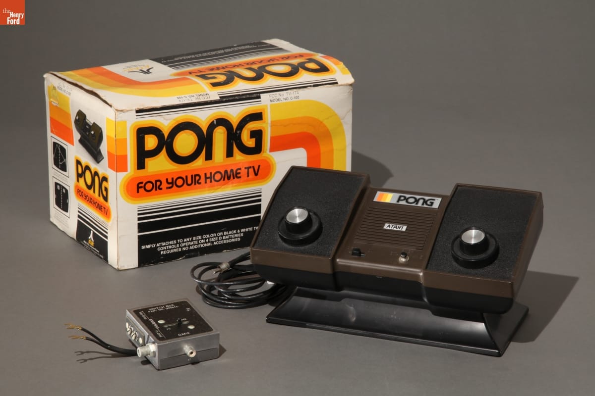 Boxy brown and black video game console with adapter and box with text and graphics in orange, yellow, and black