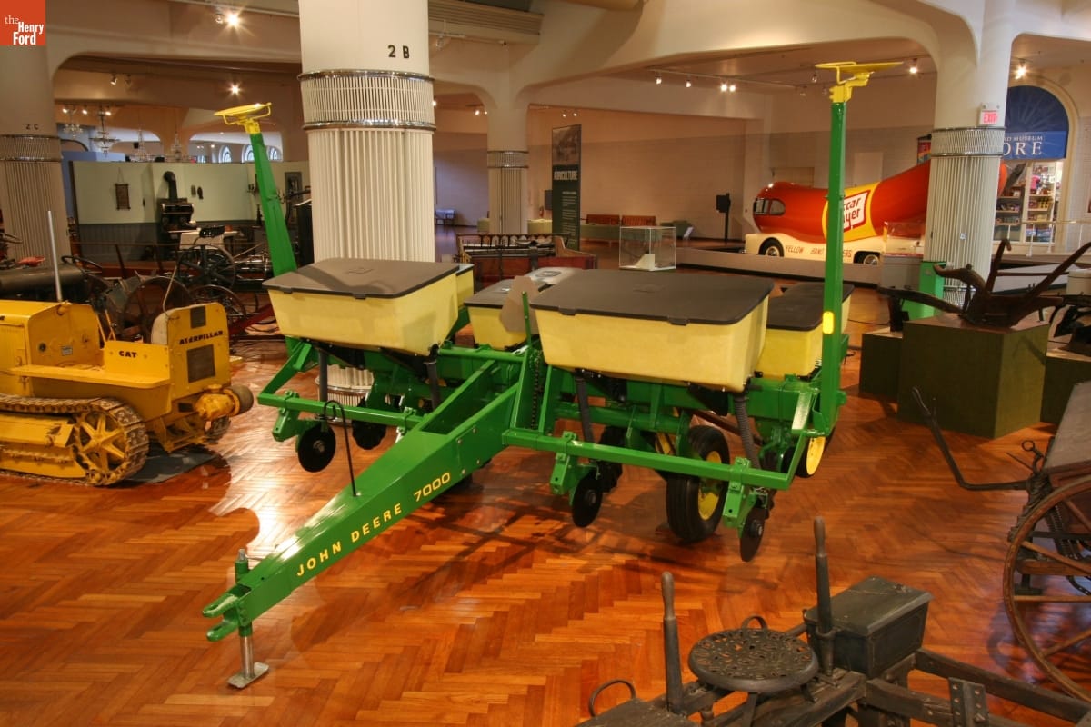 Large green agricultural implement with yellow bins, in an exhibit among other agricultural equipment and other objects