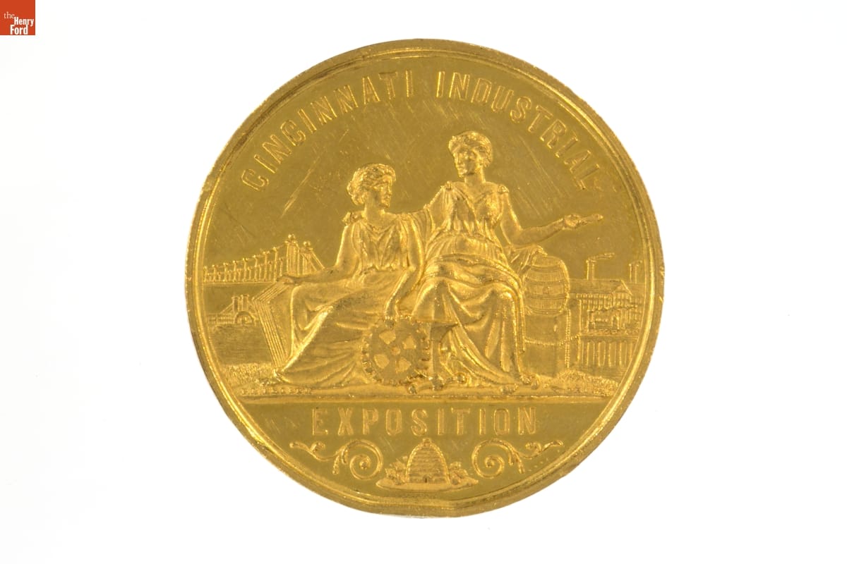 Gold coin with text and illustration of two women in classical robes with other items around them