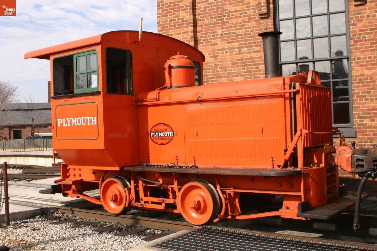 1927 Plymouth Gasoline-Mechanical Locomotive Small orange locomotive on railroad tracks by brick building