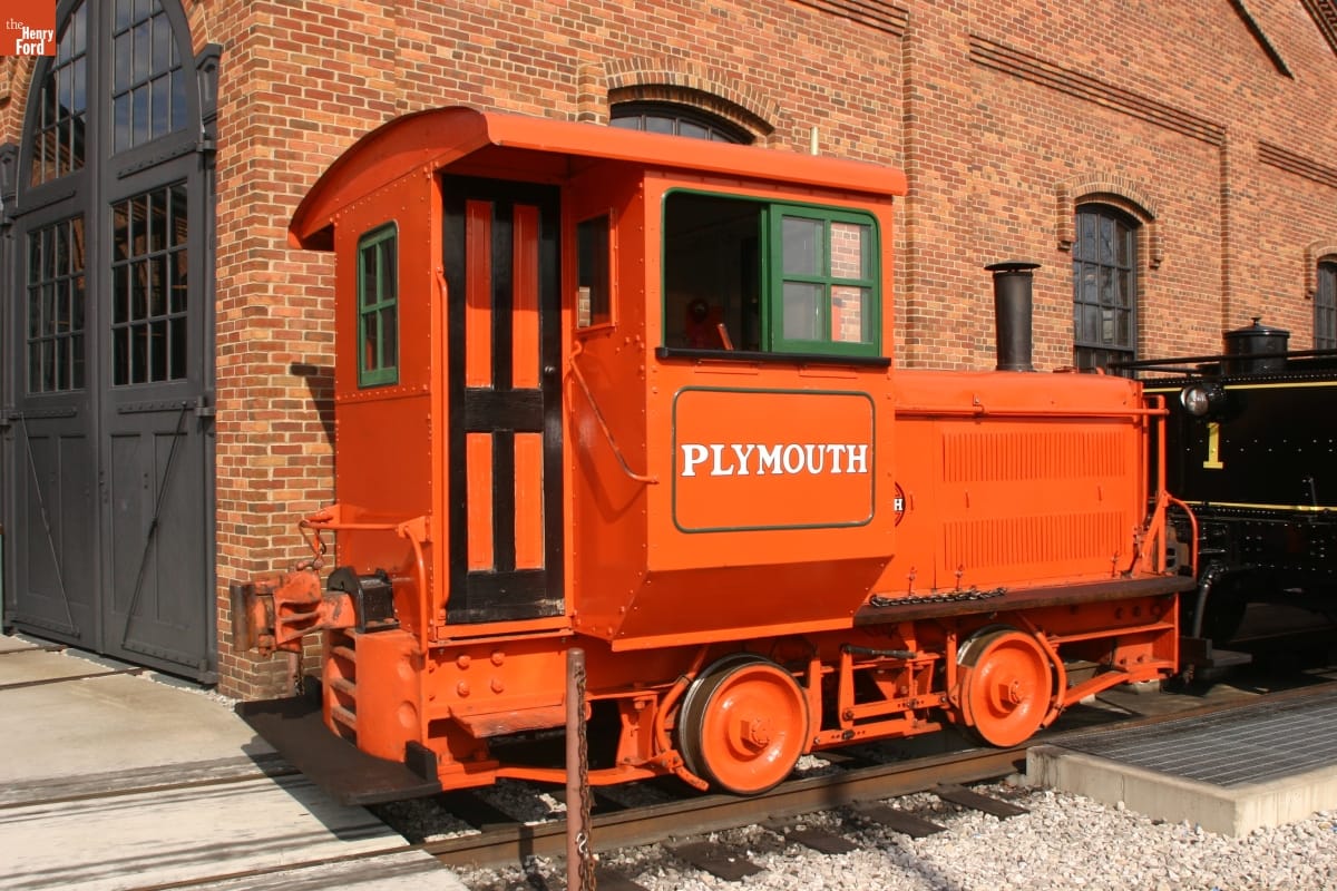 1927 Plymouth Gasoline-Mechanical Locomotive Small orange locomotive on train tracks in front of brick building