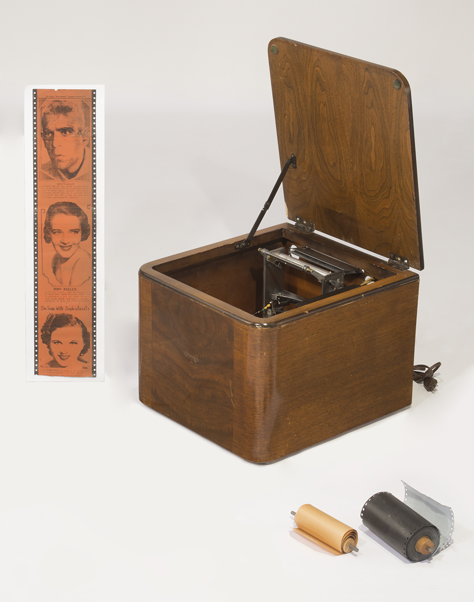 Wooden box with machinery inside, paper strip with images of three faces, paper reels
